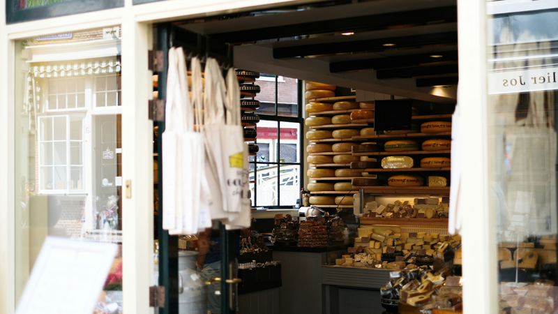Snapshot of the interior of a cheese shop. There are stacks of parmesan on the wall and totes hanging for sale along with presliced cheeses.